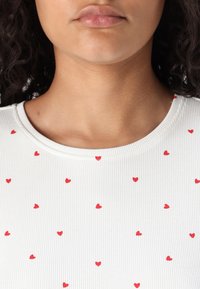 Valge ribadega kangast topp, millel on ringsuurus kaelus ja mis on kaunistatud üle pinna hajutatult paiknevate väikeste punaste südame motiividega.