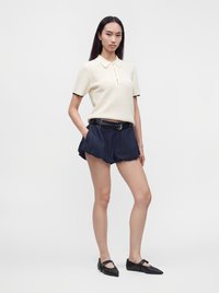 Femme debout portant un polo en maille crème à manches courtes, un short bleu marine ceinturé et des chaussures plates noires à brides.