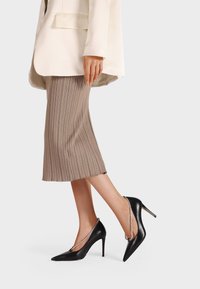 Scarpe nere in pelle con tacco alto e punta affilata, con una catena decorativa lungo il tacco. Indossate con una gonna plissettata beige.