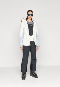 Weiße und hellblaue Skijacke mit Pelzbesatz, schwarze Skihose und schwarze Skischuhe. Blaue reflexionsverringende Skibrille rundet das Outfit ab.