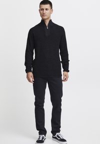 Schwarzer Strickpullover mit hohem Kragen und Reißverschluss, kombiniert mit schwarzen Jeans und schwarz-weißen Sneakern; strukturierter Stoff.