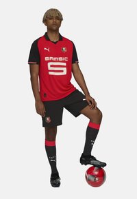 Camiseta de fútbol roja con detalles en negro, número '5', pantalones cortos negros, medias rojas y negras, y botas negras. El jugador está de pie sobre un balón de fútbol rojo.