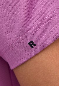 Fioletowa teksturowana tkanina z małymi perforacjami. Czarna litera "R" nadrukowana na hemie. Zbliżenie na krawędź odzieży.