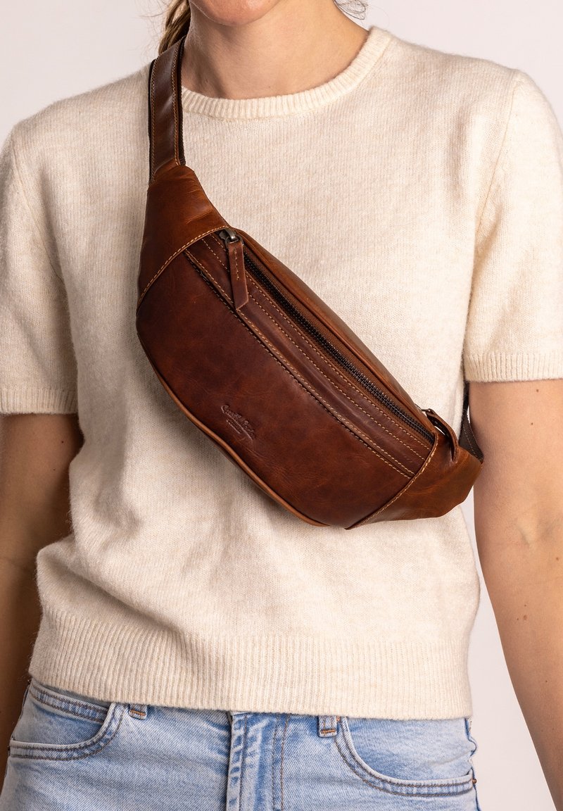 Sac bandoulière en cuir marron avec fermeture éclair porté sur un pull crème à manches courtes et un jean bleu clair sur une personne, visible des épaules à la taille.