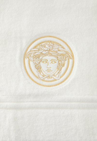 Λευκή πετσέτα με περίπλοκη χρυσή κεντητική ενός κεφαλιού Μέδουσας μέσα σε κυκλικό σχέδιο, που αναδεικνύει πολυτελή υφή και λεπτομέρεια.