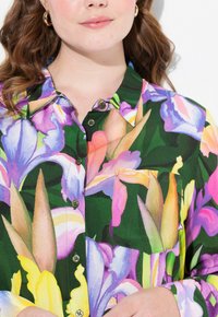 Blusa floreale con sfondo verde decorato da grandi fiori viola e gialli, design con colletto e bottoni davanti, comprensiva di taschino sul petto.