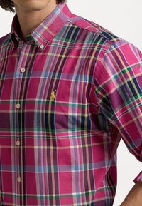 Rutig rosa skjorta med knappar som har olika ränder i lila, grönt och gult. Har en knappad krage och en liten gul logotyp på bröstet.