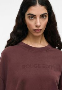 Bordo spalvos džemperis su apvaliu kaklu, su subtiliu iškilniu tekstu "ROUGE EDIT" priekyje. Minkšta medžiaga, laisvas siluetas.
