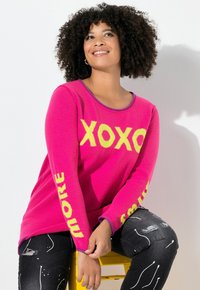 Frau mit lockigem Haar, die einen leuchtend rosa Pullover mit gelbem "XOXO" auf der Vorderseite und Text an den Ärmeln trägt, sitzt auf einem gelben Hocker vor einem weißen Hintergrund.