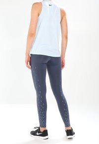 Top sin mangas deportivo azul combinado con leggins oscuros que presentan rayas verticales blancas. El atuendo se completa con zapatillas deportivas negras.