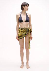 Čierny vrchný diel bikín so šnurovaním za krkom a zlatým kruhom, doplnený žltým vzorovaným sarongom zviazaným v páse, ktorý sa nosí cez boky.