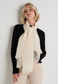 Bufanda de color crema con tela texturizada y acanalada. Colocada de manera casual alrededor del cuello, presenta bordes suaves y un diseño en capas.