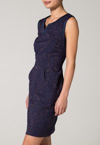 Tmavě modré krajkové šaty s přiléhavým střihem, bez rukávů a jemnými květinovými vzory. Mají výstřih do V a nařasený pas.