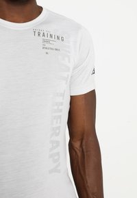 Vit tränings t-shirt med korta ärmar, med svart text på framsidan: "TRAINING" och "SWEAT THERAPY," med en logotyp på ärmen.