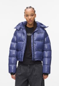 Marineblauwe pufferjack met een glanzende afwerking, voorzien van een capuchon, ritssluiting en drukknoopsluitingen. Draag het over een zwart shirt en een grijze broek.