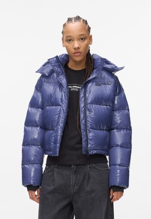 Marineblauwe pufferjack met een glanzende afwerking, voorzien van een capuchon, ritssluiting en drukknoopsluitingen. Draag het over een zwart shirt en een grijze broek.