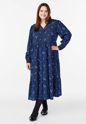 Frau in einem blauen, langärmeligen, gestuften Midikleid mit kleinen Blumenmustern, schwarzen Strumpfhosen und flachen schwarzen Schuhen, die mit einer Hand auf der Hüfte steht.