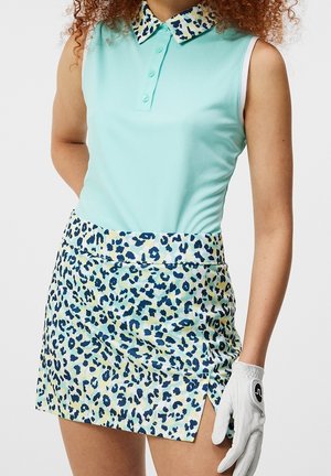 Kobieta w bezrękawniku w kolorze aqua polo i niebieskiej spódnicy do golfa w kropki, trzymająca białą rękawiczkę golfową na prawej dłoni.