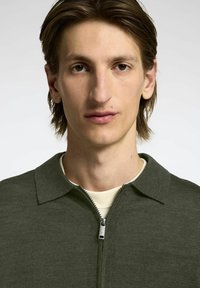 Tamsiai žalia zip-up megztinis su apykakle, pagamintas iš tekstūruoto megztinio audinio. Yra sidabrinė užtrauktukas ir lengvas pamušalas.