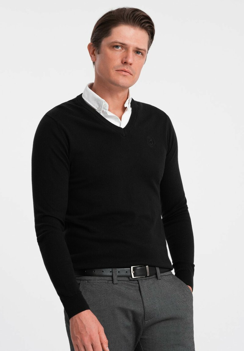 Schwarzer V-Ausschnitt-Pullover aus weichem Material, mit langen Ärmeln und einem dezenten Logo auf der Brust, kombiniert mit grauen strukturierten Stoffhosen und einem schwarzen Gürtel.