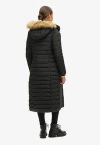 Abrigo de plumas negro con un diseño largo y ajustado, textura acolchada vertical y capucha forrada de pelo sintético. Llevado con pantalones y botas negras.