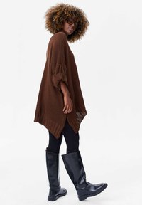Bruine gebreide oversized poncho met ribbeltextuur, zijsplitsingen, gecombineerd met zwarte leren laarzen tot de knie en zwarte leggings.