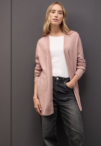 Ljusrosa cardigan med ribbade kanter, öppen design framtill, långa ärmar, bärs över en vit topp och kombineras med mörka jeans. Slät textur.