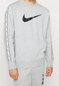 Grå sweatshirt med rund hals, svart Nike-logotyp på bröstet, och vita accentlinjer längs ärmarna med svarta logotyper. Mjuk texturerad tyg.
