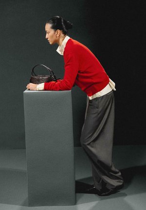 Czerwony sweter na białej koszuli, szare spodnie o szerokich nogawkach oraz teksturowana ciemna torebka na szarym pedestale na ciemnym tle.