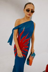 Donna che indossa un vestito blu a spalla unica con un grande design sun rosso, occhiali da sole marroni, e tiene una pochette blu abbinata con motivo a sole.