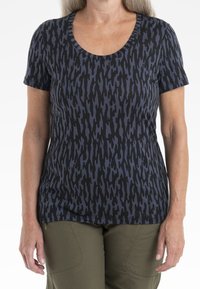 Kvinna iförd en kortärmad skjorta med svart och mörkblå abstrakt mönster och olivgröna byxor, stående med avslappnade händer vid sidorna.