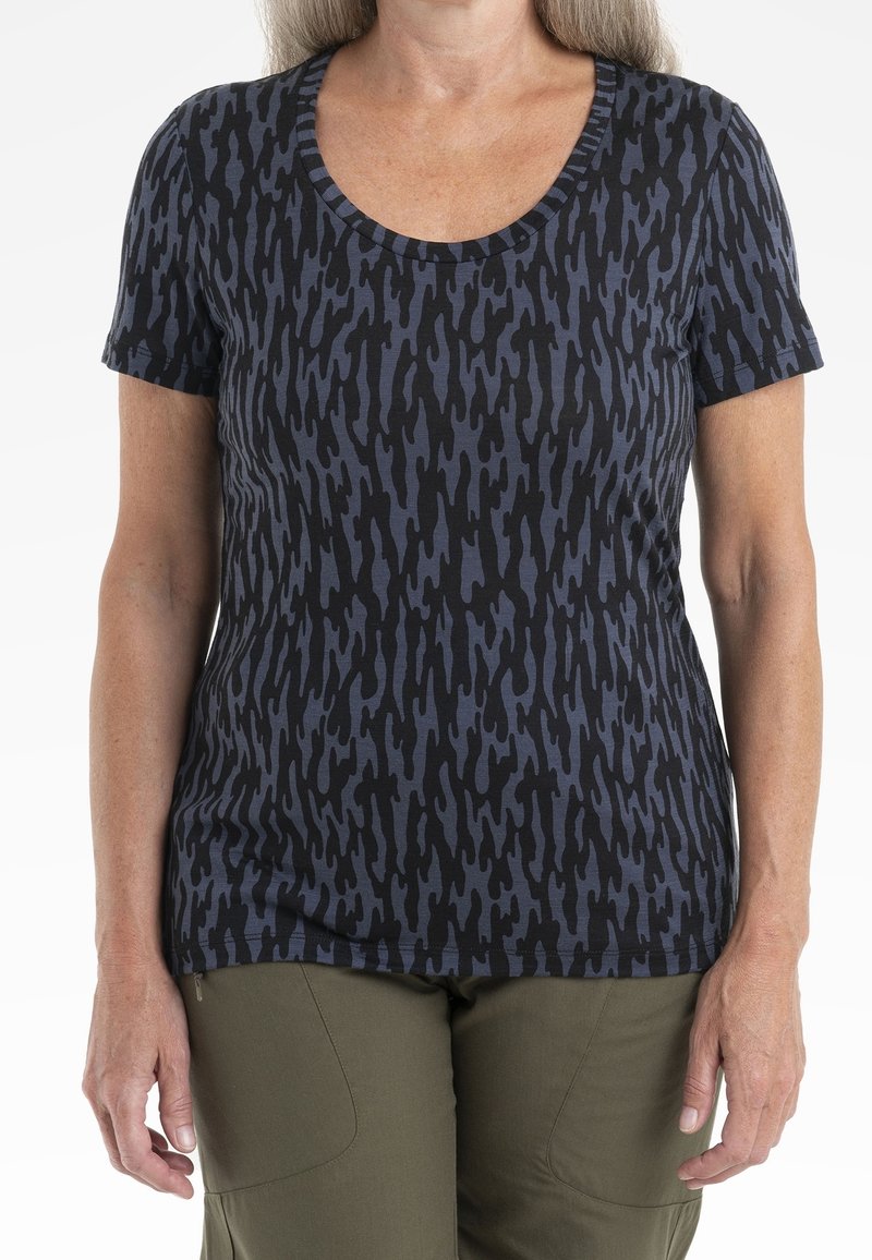 Kvinna iförd en kortärmad skjorta med svart och mörkblå abstrakt mönster och olivgröna byxor, stående med avslappnade händer vid sidorna.