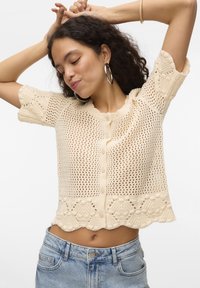 Cardigan en tricot à manches courtes, de couleur crème, avec une maille ouverte, un col rond et une fermeture à boutons. Présente un ourlet à volants et des détails texturés.