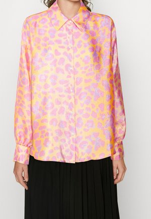 Kobieta w koszuli na długi rękaw z błyszczącym różowo-pomarańczowym wzorem w cętki i czarnej plisowanej spódnicy.