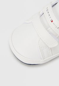 Λευκό συνθετικό παπούτσι με λουράκια βέλκρο, στρογγυλεμένη μύτη, αντίθετη μπλε σόλα και εμφανές λογότυπο «TOMMY HILFIGER» στο λουράκι.