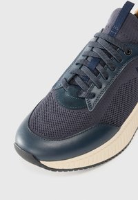 Tênis azul-marinho com tecido em malha e detalhes em couro liso, apresentando uma sola texturizada, bico redondo e cadarços planos.