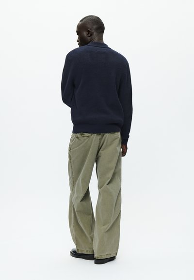 Mornarsko pleteni pulover in ohlapne svetlo olivno zelene hlače. Pulover ima teksturiran vzorec, medtem ko imajo hlače ohlapen kroj in vidne šive.