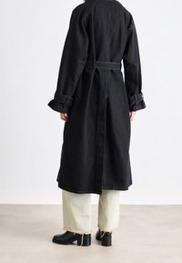 Lungo cappotto di denim nero con vita con cintura, polsini e spacco posteriore. Abbinato a pantaloni verde chiaro con orlo sfrangiato e stivaletti neri.
