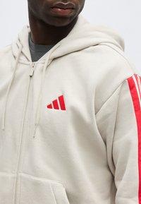 Homme portant un sweat à capuche beige clair avec fermeture éclair, orné d'un logo à rayures rouges sur la poitrine et de rayures rouges le long de la manche, tourné légèrement vers la gauche.