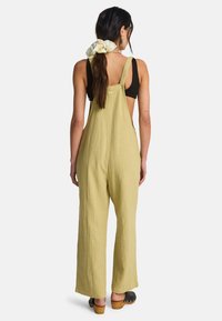 Φωτεινά χακί λιναρά ολόσωμα παντελόνια με φαρδιά πόδια και ρυθμιζόμενους ιμάντες, με ανοιχτή πλάτη και συνδυασμένα με μαύρο τοπ και ξύλινα τακούνια.