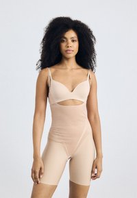 Beige bodysuit, jossa on leikattu yksityiskohta, säädettävät olkaimet ja tiukka malli, valmistettu sileästä materiaalista saumatonta viimeistelyä varten.