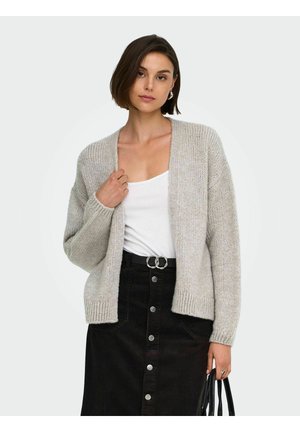 Femme portant un cardigan en tricot gris clair par-dessus un débardeur blanc, avec une jupe noire à boutons devant, tenant un sac noir d'une main.