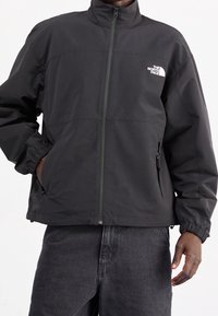 Chaqueta cortavientos negra con cuello alto, cierre de cremallera, puños elásticos y bolsillos laterales. Presenta un logo blanco en la parte superior izquierda del pecho.