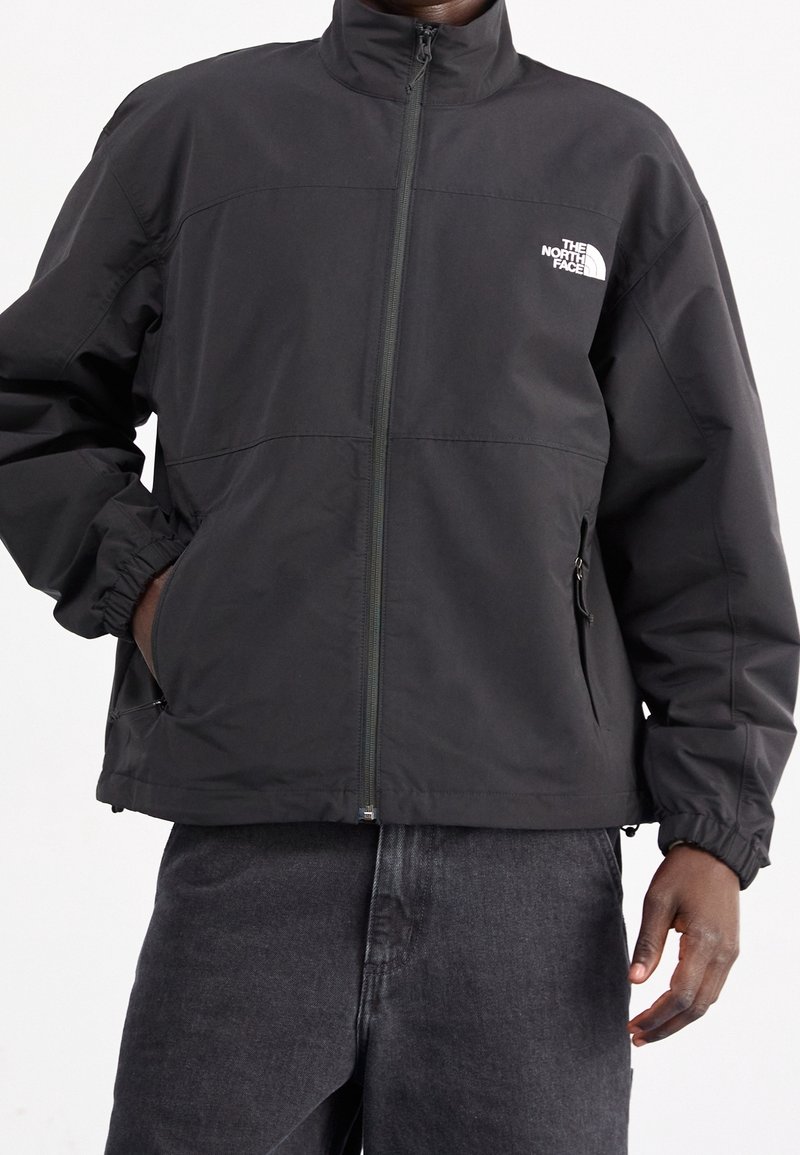 Chaqueta cortavientos negra con cuello alto, cierre de cremallera, puños elásticos y bolsillos laterales. Presenta un logo blanco en la parte superior izquierda del pecho.