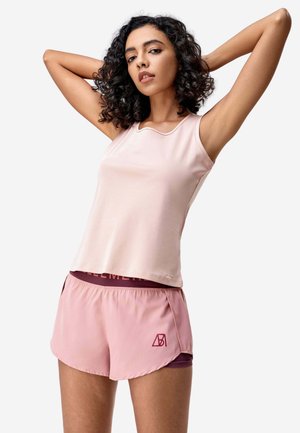 Donna con capelli ricci che indossa un top smanicato rosa chiaro e pantaloncini sportivi abbinati con un logo, posa con le braccia sollevate dietro la testa.