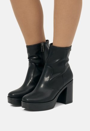 Botas negras hasta el tobillo hechas de cuero suave con puntera redonda, cremallera lateral y un tacón alto y robusto de textura estriada.