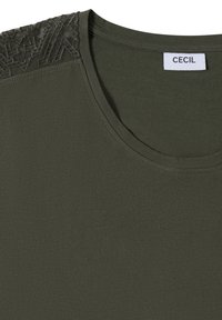 T-shirt vert olive foncé à col rond avec un détail en dentelle noire texturée sur l'épaule gauche et une étiquette blanche portant la mention « CECIL » à l'intérieur du col.