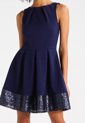 Granatowa sukienka bez rękawów z plisowaną spódnicą oraz szeroką lamówką z ciemnoniebieskich cekinów, noszona na gołych ramionach i nogach.