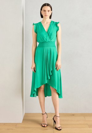 Grünes, fließendes Kleid mit Rüschenschultern und gesmoktem Bund, das einen asymmetrischen Saum und eine weiche Drapierung aufweist. Schuhe: schwarze, geschnürte High Heels.
