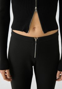 Černý žebrovaný pletený krátk�ý zipový top s dlouhými rukávy, v kombinaci s černými elastickými kalhotami s viditelným zipovým detailem.